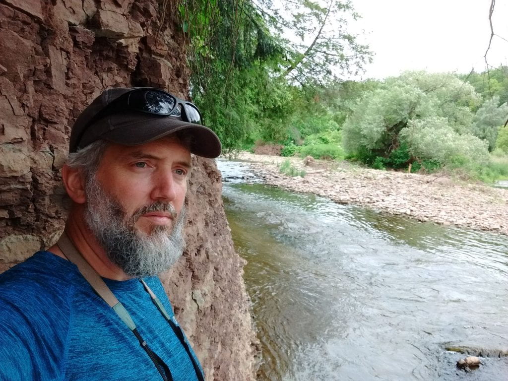 man at the edge of a river with a rock face behind him